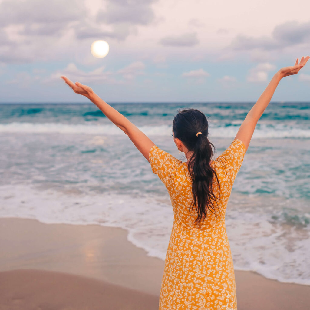 Woman enjoying sunset with full moon on the beach July 2023